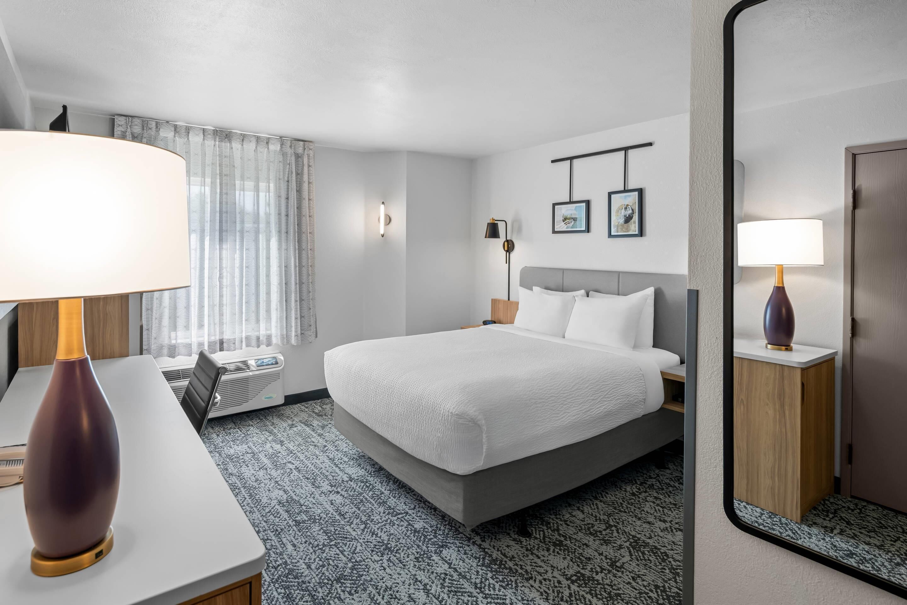 Una habitación de hotel con cama, escritorio con lámpara, espejo, un cuadro en la pared y ventana con cortinas.