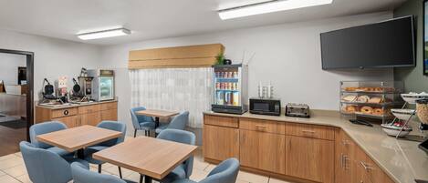 A modern kitchen with a flat-screen TV, a microwave, and a coffee machine. There are blue chairs around a wooden table, and a buffet counter with baked goods.