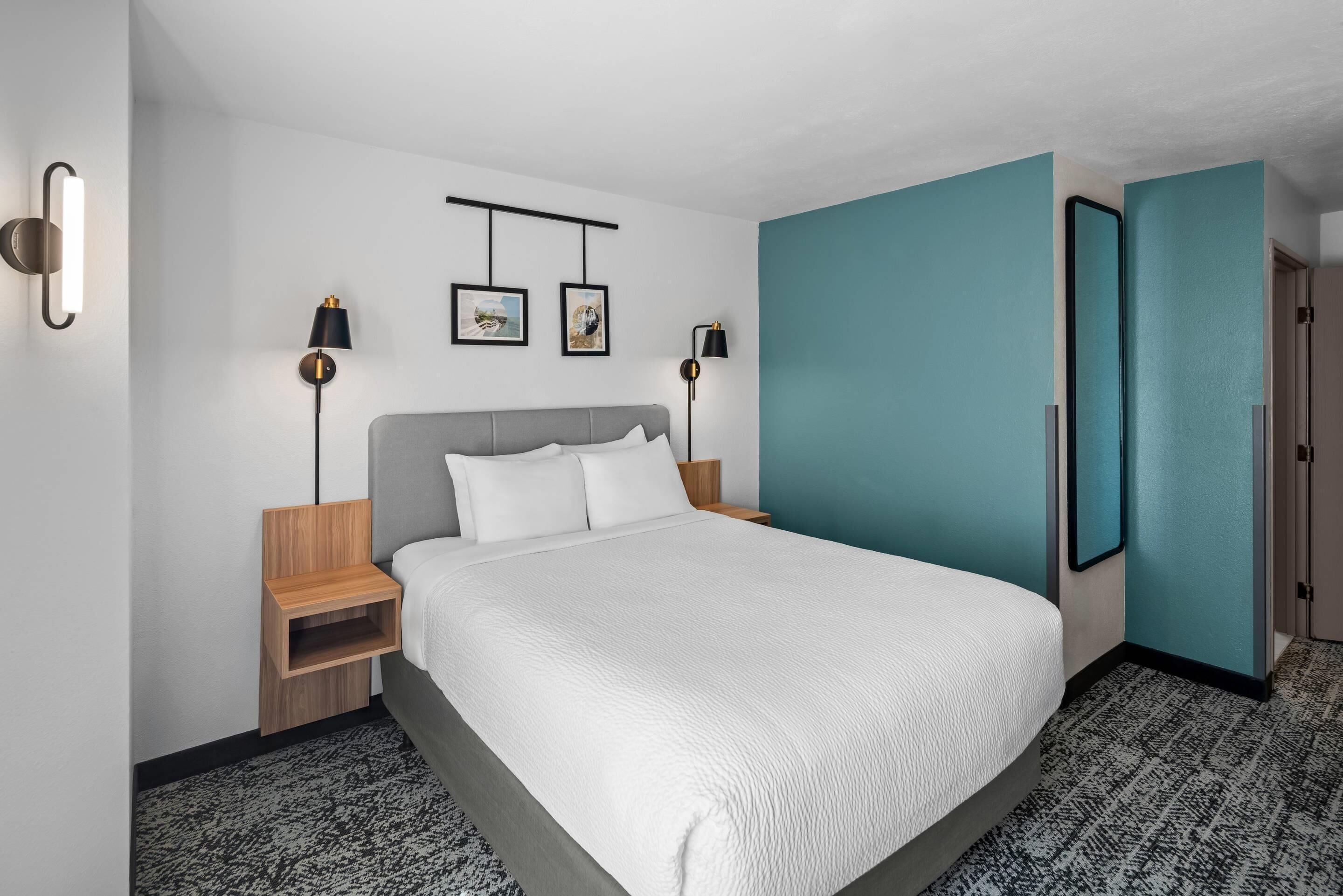 A modern hotel room with a large bed, two bedside tables, a nightstand, a mirror, and artwork on the wall.
