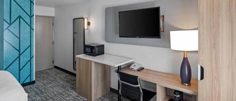 A hotel room with a desk, chair, TV, and a minibar.
