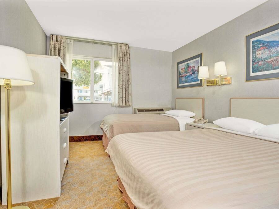 A hotel room with two beds, a desk, a lamp, a window with curtains, and wall-mounted lamps.