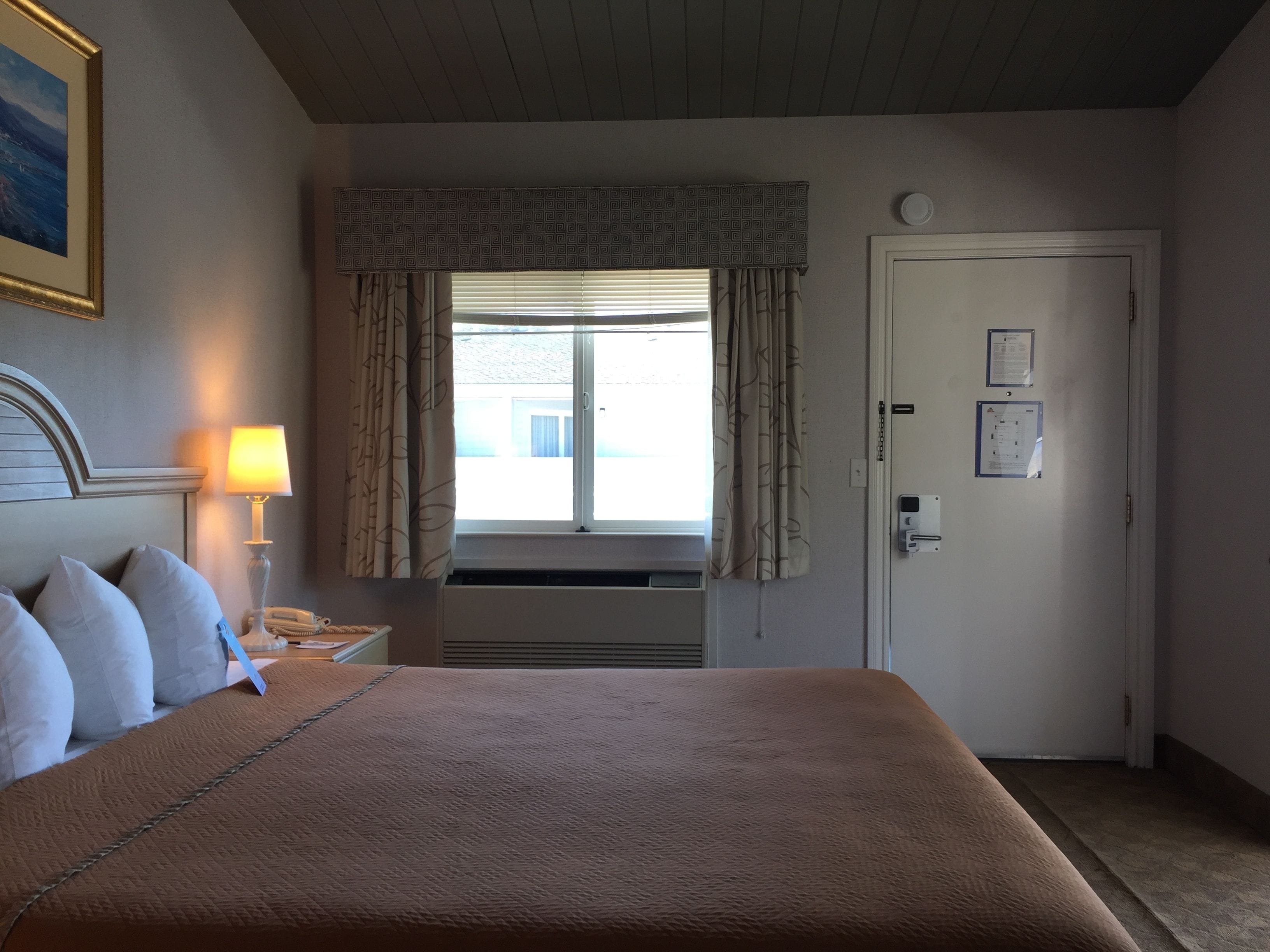 Habitación de hotel con una cama grande, una lámpara de noche, una ventana con cortinas, una puerta con cerradura y un aire acondicionado instalado en la pared.