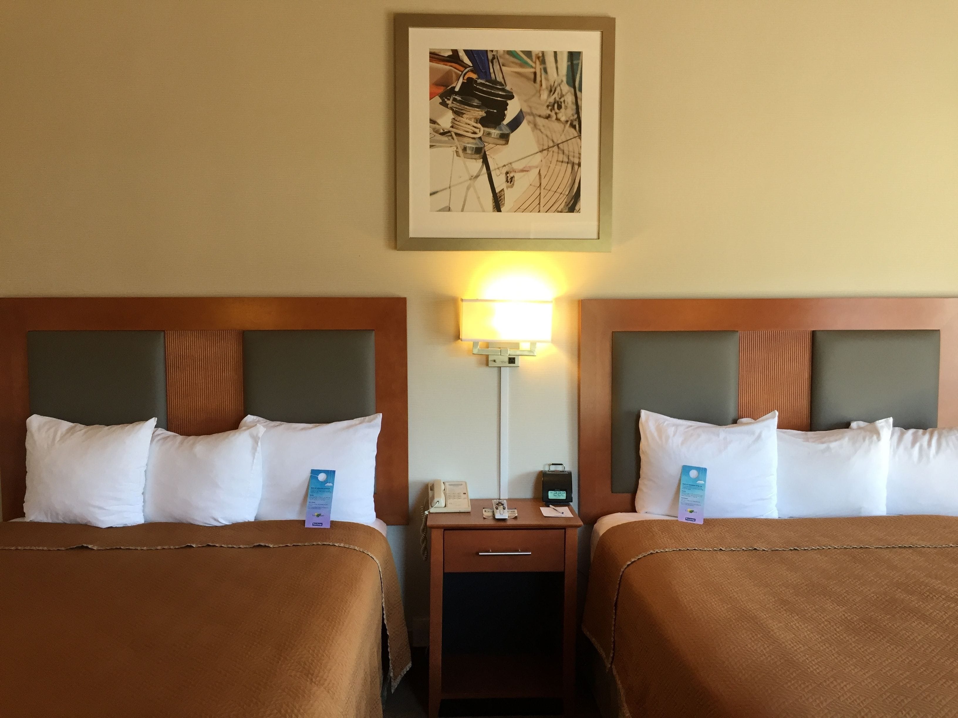 Quarto de hotel com duas camas, cada uma com cabeceira marrom, roupa de cama branca e mesa de cabeceira com abajur e telefone.