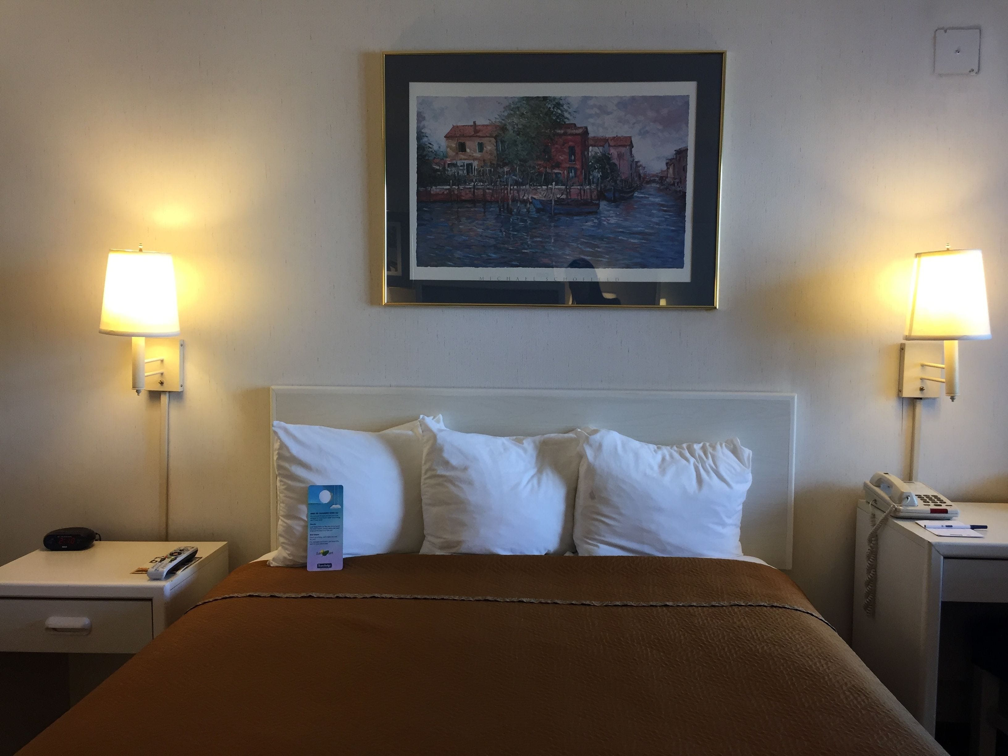 Habitación de hotel con cama, dos lámparas de noche, un cuadro enmarcado con una escena de canal y una mesita de noche con teléfono y control remoto.