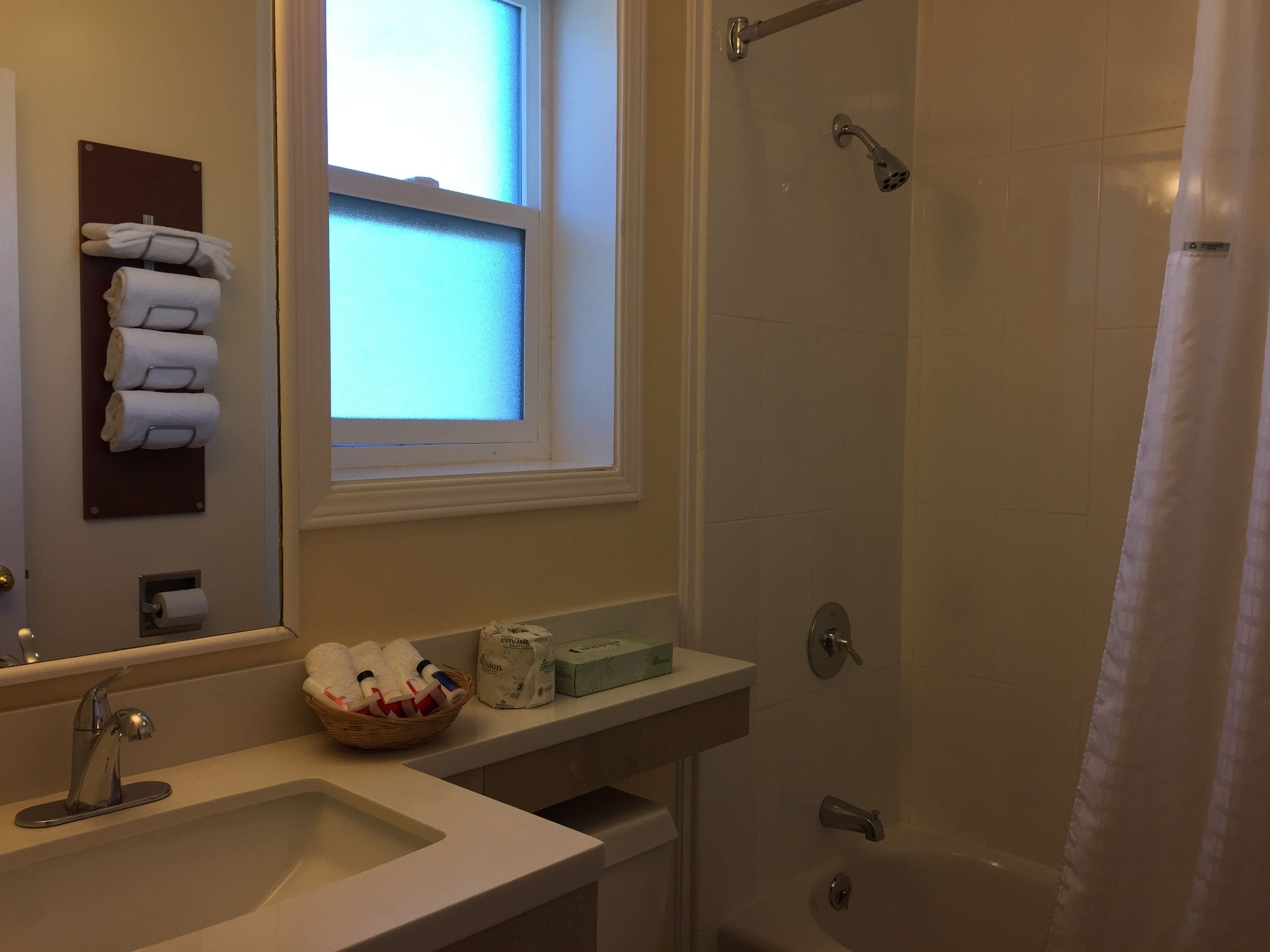 A bathroom with a sink, mirror, towel rack, shower, and window.