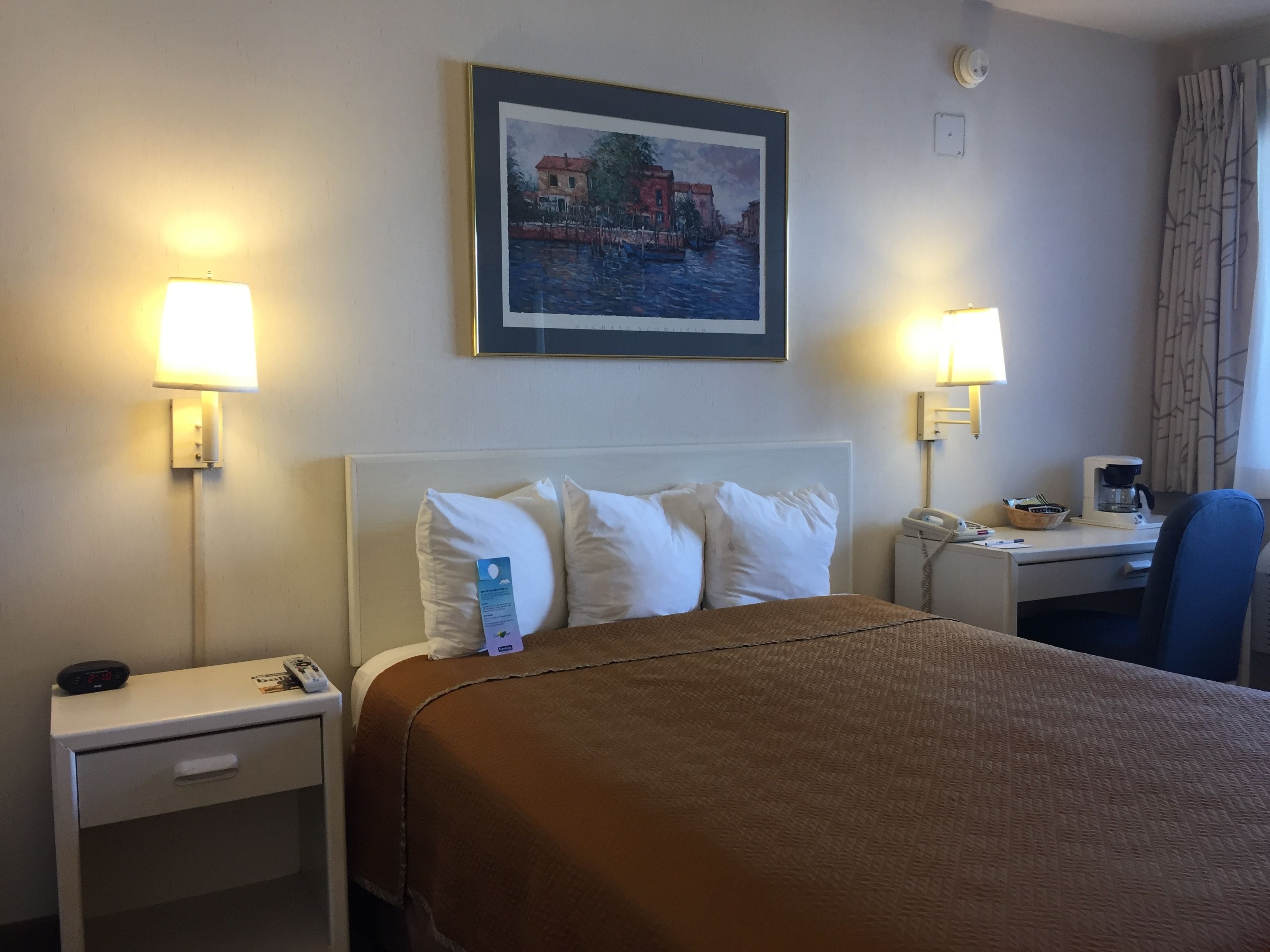 A hotel room with a bed, two bedside tables, a desk with a chair, a painting on the wall, and two lamps.