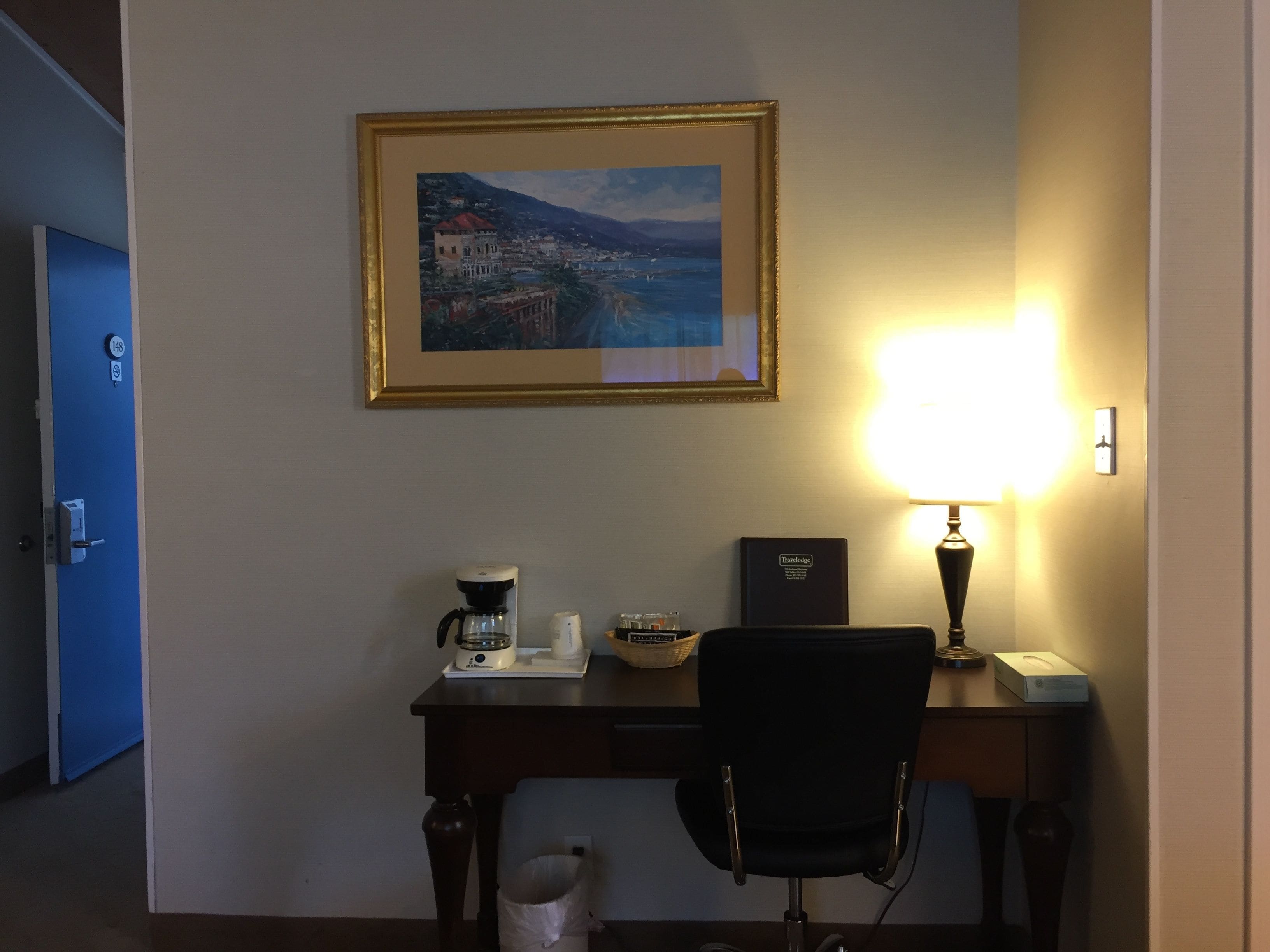 A hotel room with a desk, a coffee maker, a lamp, a framed picture of a coastal town, and a trash bin.