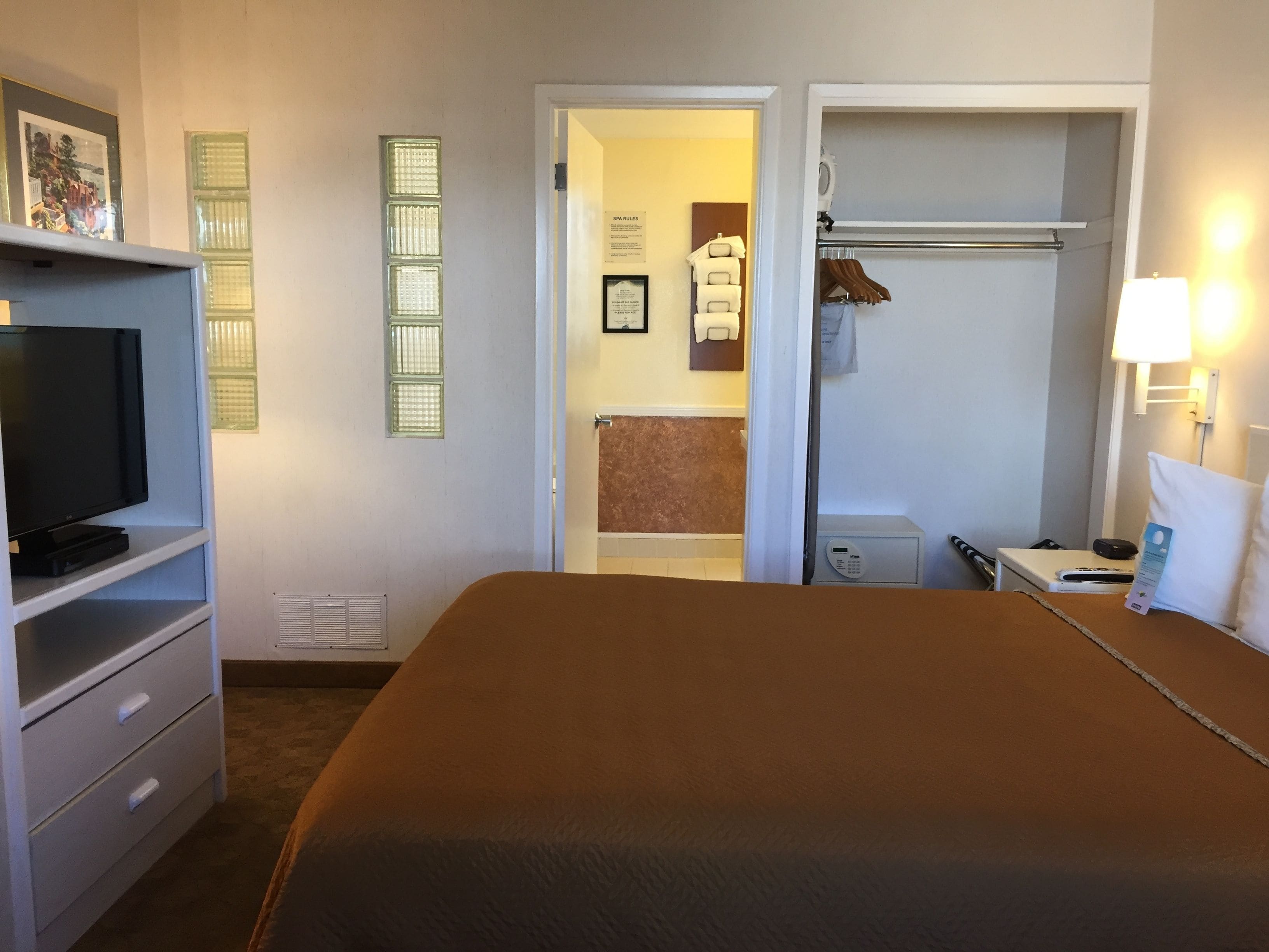 A hotel room with a bed, a TV on a stand, a bathroom visible through an open door, and a coat rack with a towel.