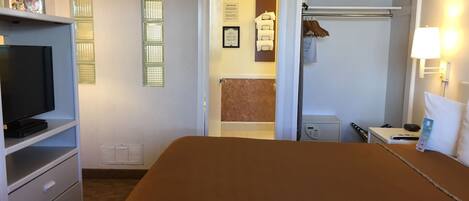 A hotel room with a bed, a TV on a stand, a bathroom visible through an open door, and a coat rack with a towel.