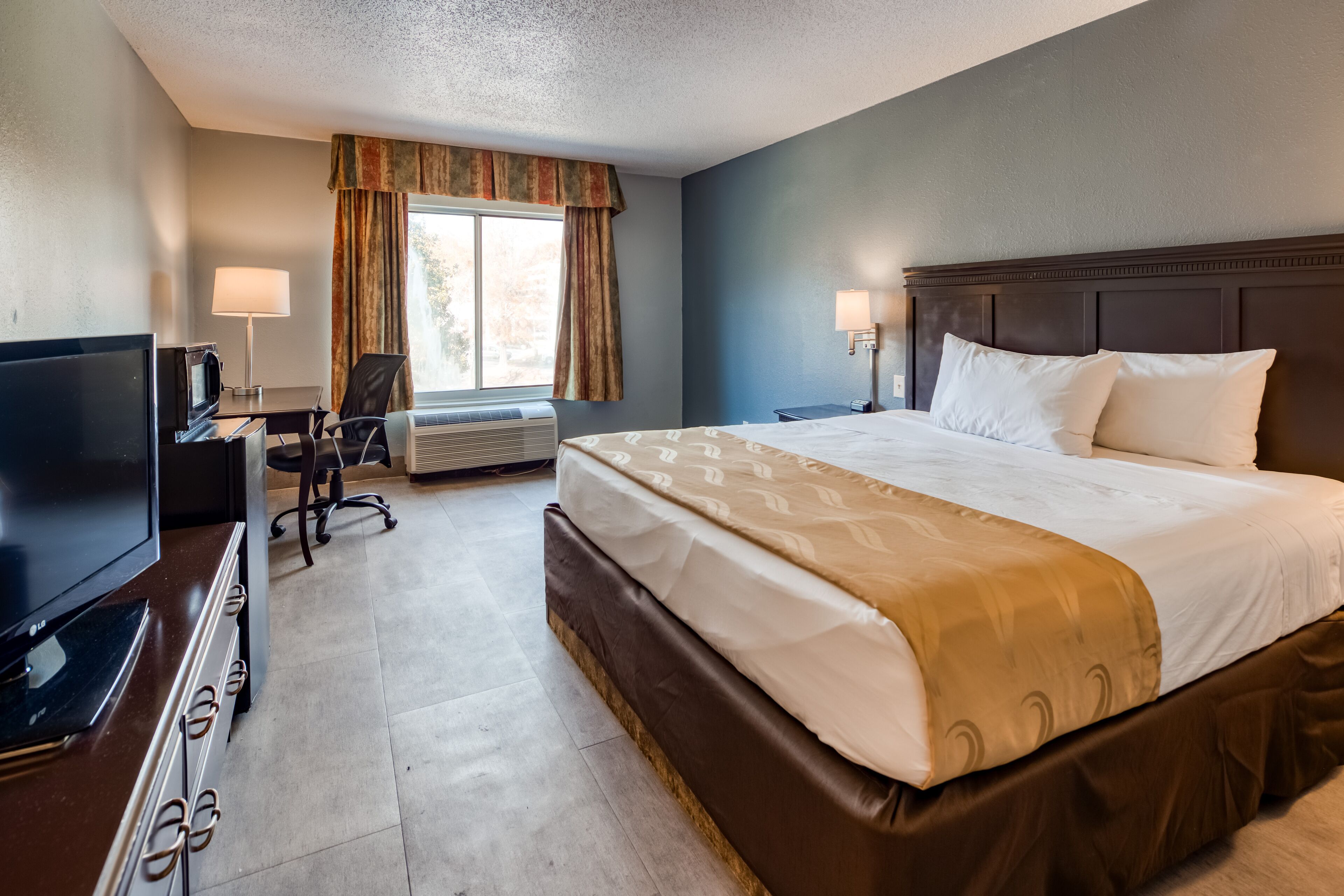 Quarto de hotel com uma cama grande, uma mesa de trabalho com televisão e uma cadeira.