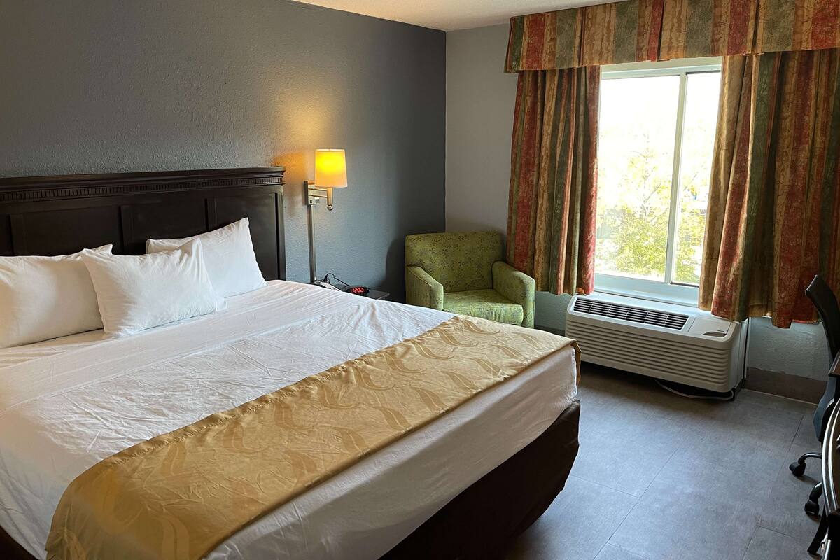 Een hotelkamer met een groot bed, een nachtkastje met een lamp, een groene fauteuil en een raam met gordijnen.