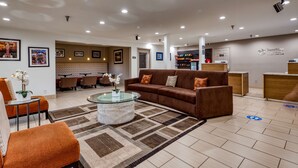 Eine Hotelhalle mit einem braunen Sofa, einem Glastisch und einem gemusterten Teppich. An den Wänden hängen Bilderrahmen, und im Hintergrund ist eine Rezeption erkennbar.
