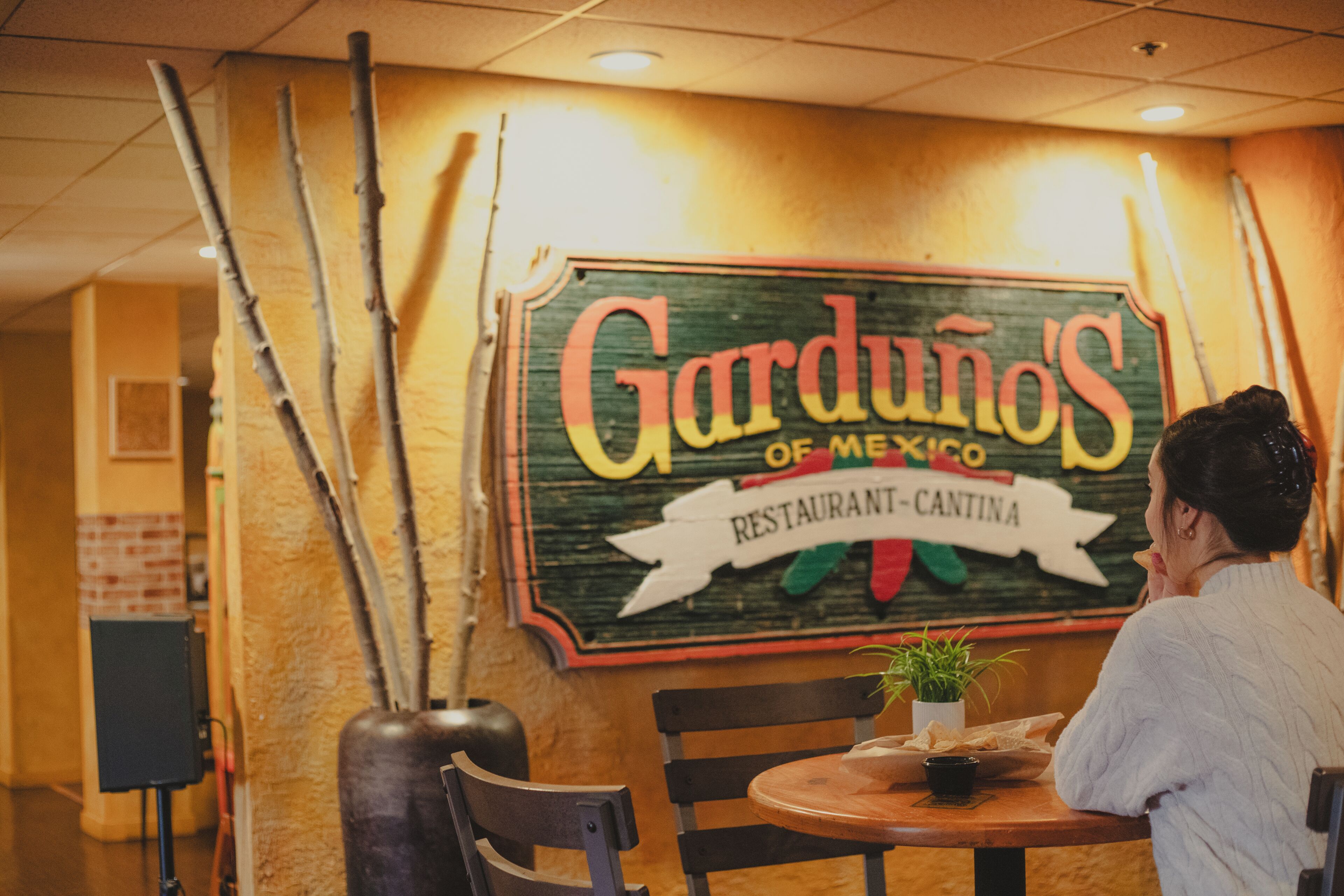 A restaurant interior with a sign reading 'Garduinos of Mexico Restaurant-Cantina,' a wooden table, chairs, and a potted plant.