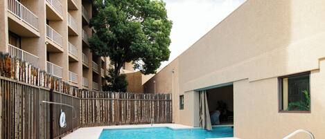 A swimming pool with a wooden fence, a building with balconies, and a tree in the background.