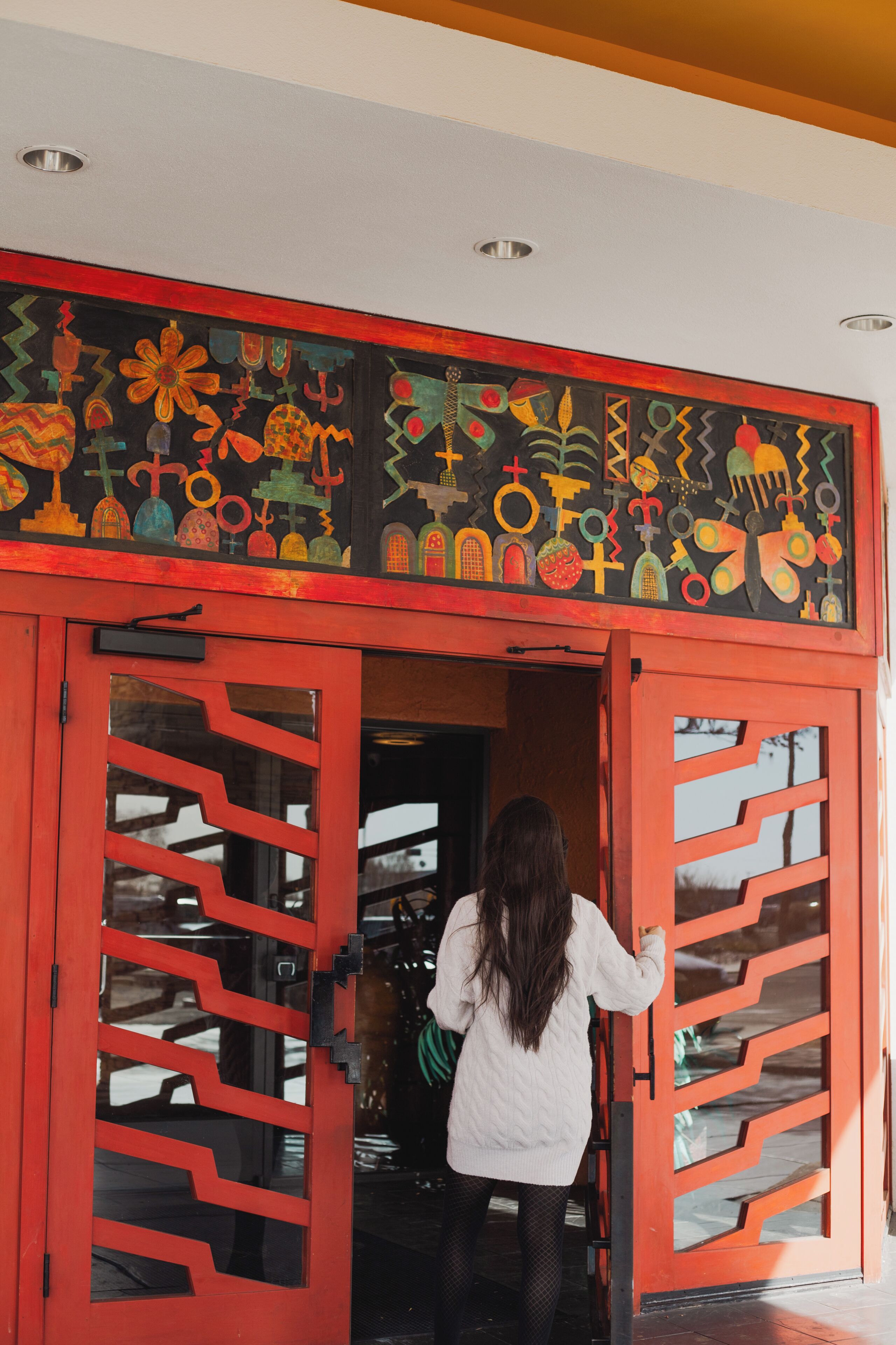 A woman entering a building through a red door with a decorative black and red mural above it.