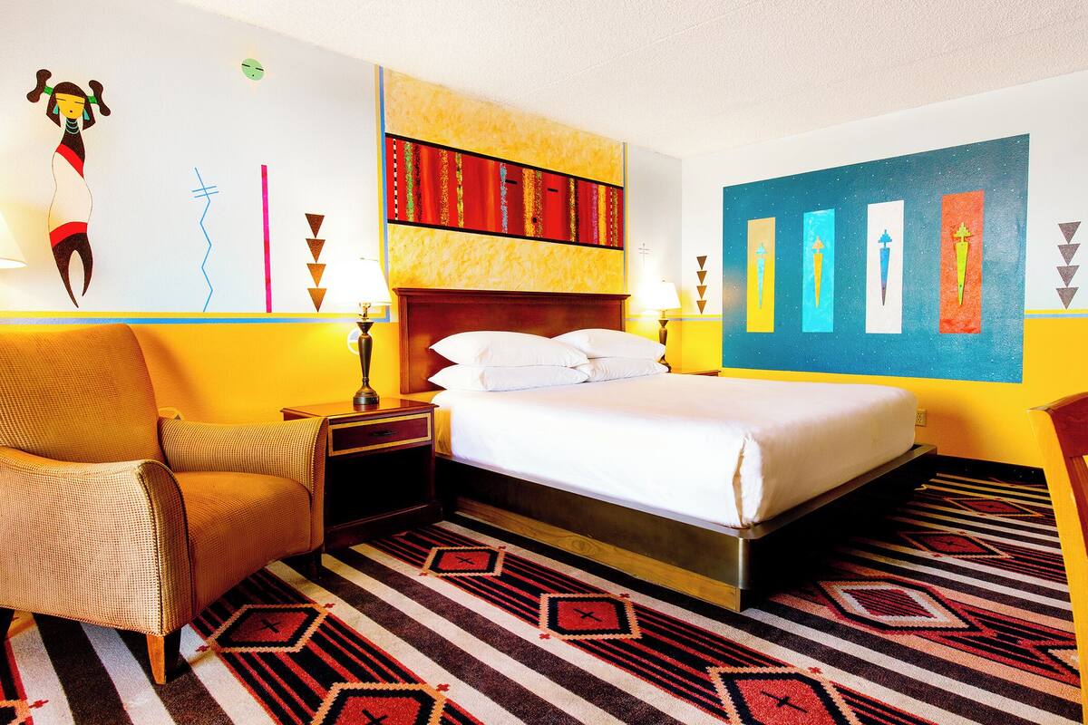 Habitación de hotel con cama, mesitas de noche, una silla, un escritorio de madera y una pared con arte colorido.