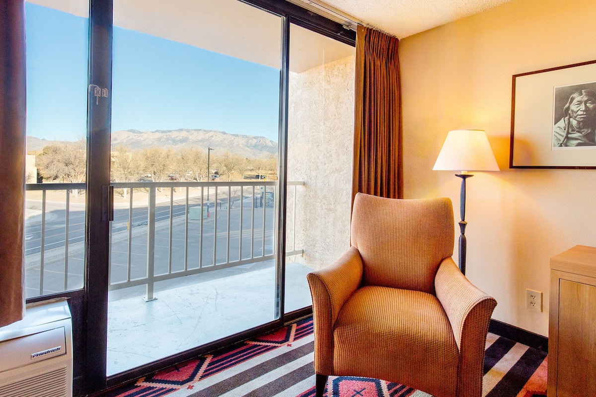 Habitación de hotel con un ventanal grande, una silla, una lámpara y un cuadro en la pared.