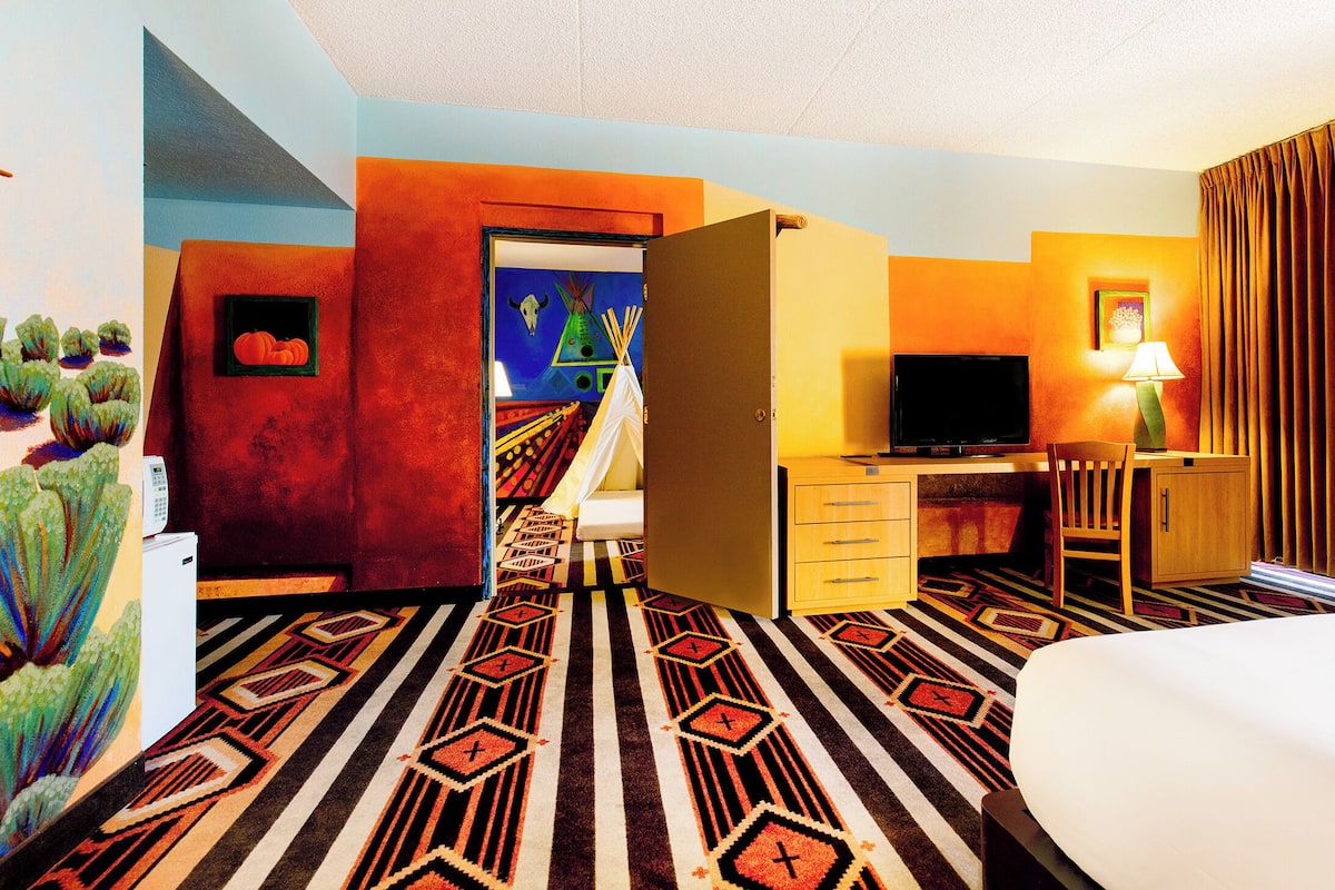 Habitación de hotel con cama, escritorio con televisor, silla, lámpara y puerta que da a un pasillo con un cuadro.