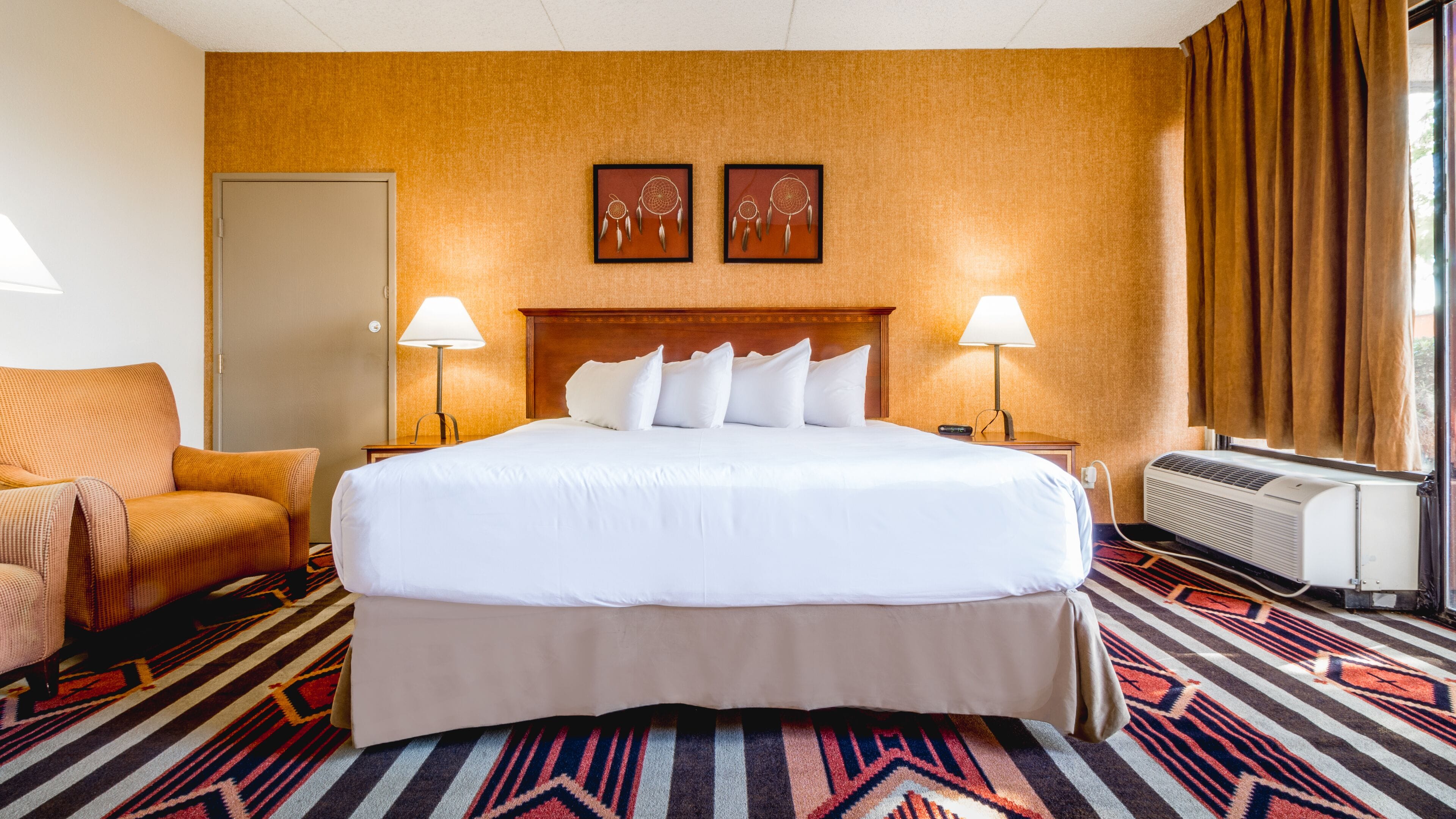 Une chambre d’hôtel avec un grand lit, deux lampes de chevet, un climatiseur fixé au mur, une moquette à motifs et une porte.