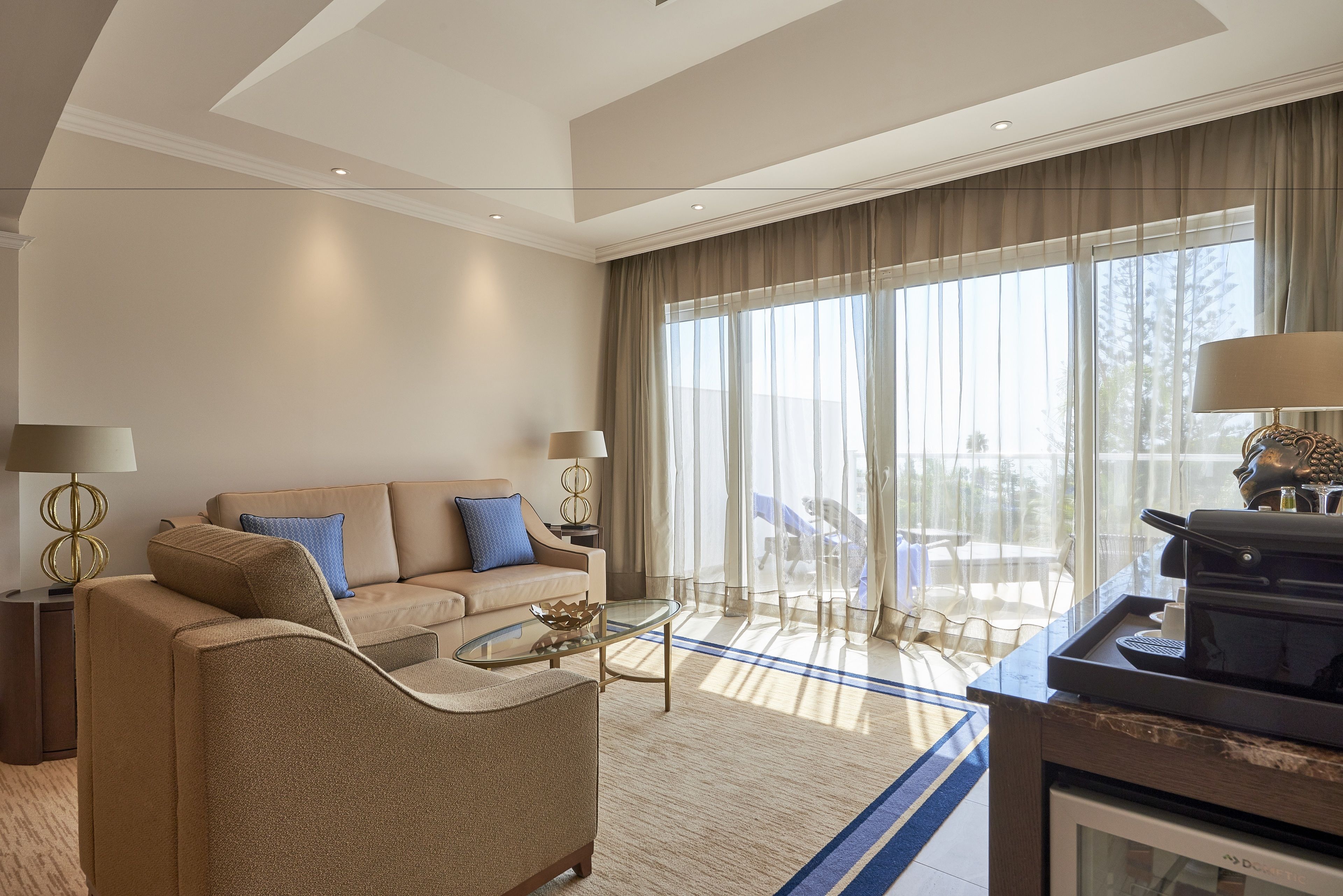 A modern hotel room with a beige sofa, a glass coffee table, and a large window with sheer curtains.