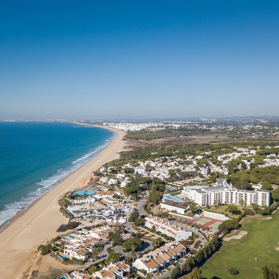 An aerial view of a coastal resort with beachfront villas, a golf course, and a large hotel.