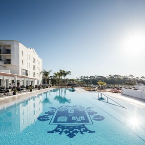 A swimming pool with a decorative tile pattern, surrounded by lounge chairs and umbrellas, with a building and palm trees in the background. - Dona Filipa Hotel (Loulé)