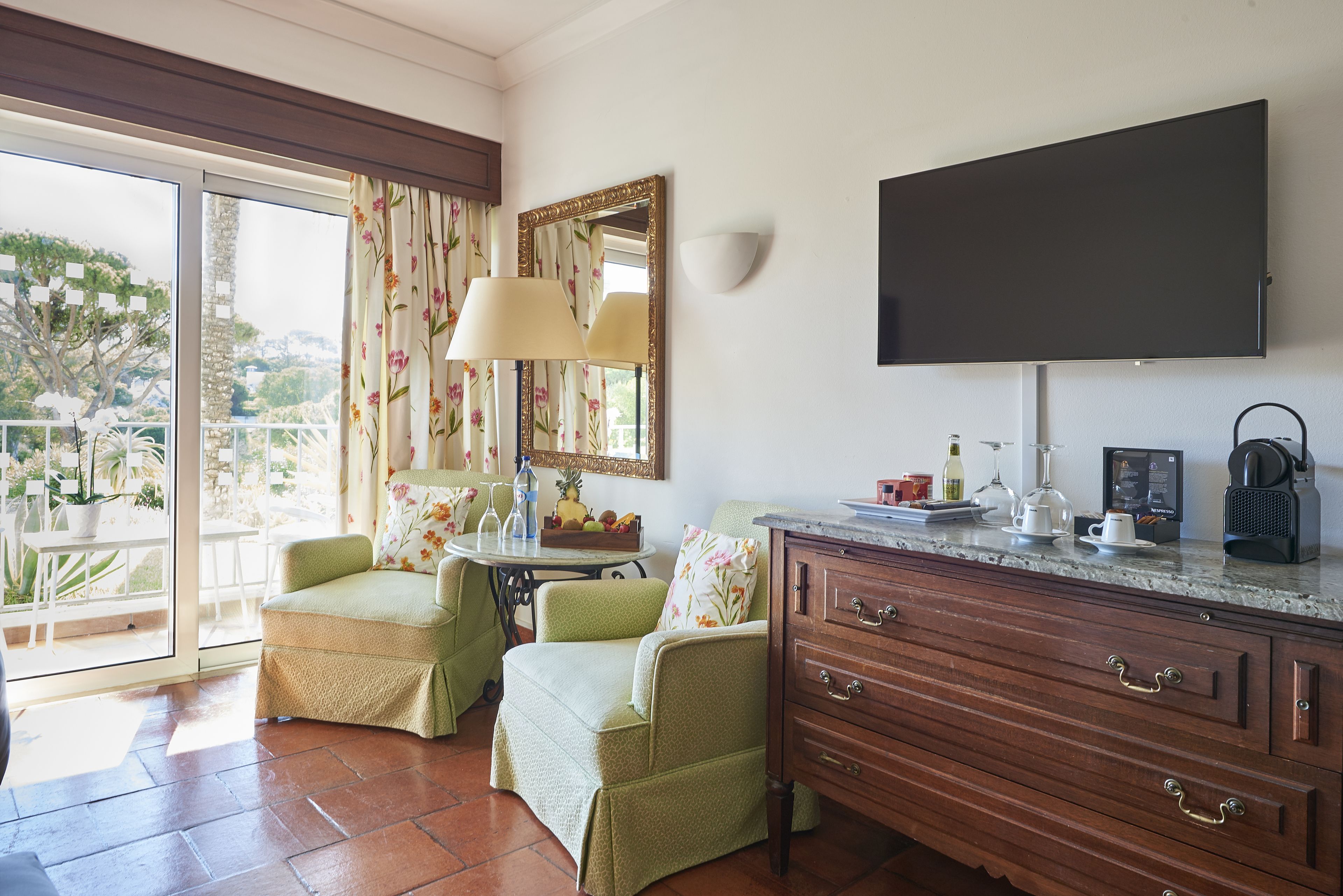 A room with a TV, wooden cabinet, two green armchairs, a table with a lamp, and a balcony with a view.