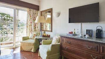A room with a TV, wooden cabinet, two green armchairs, a table with a lamp, and a balcony with a view.