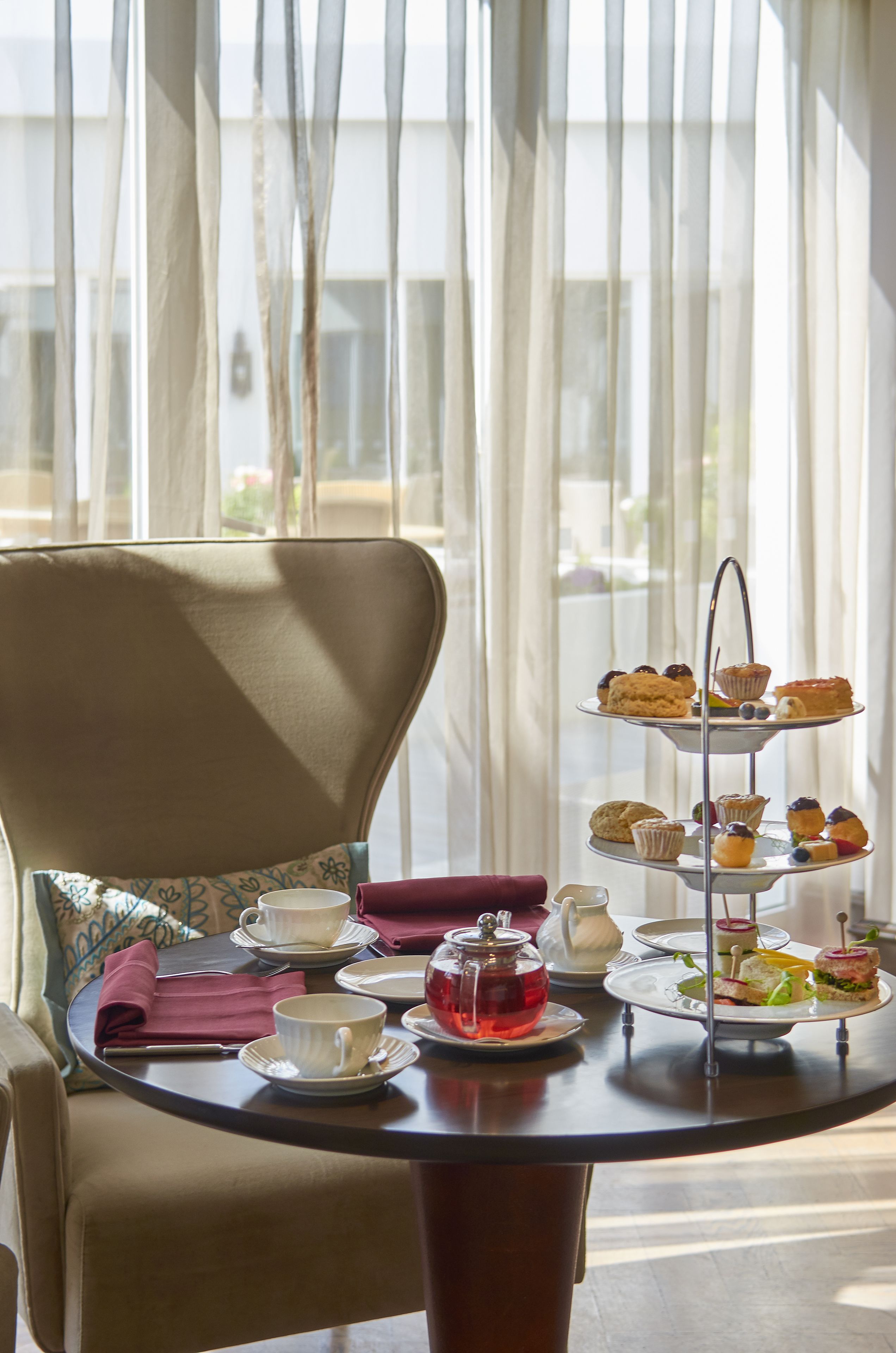A round table with a tea set, a three-tiered dessert stand, and a chair with a cushion.