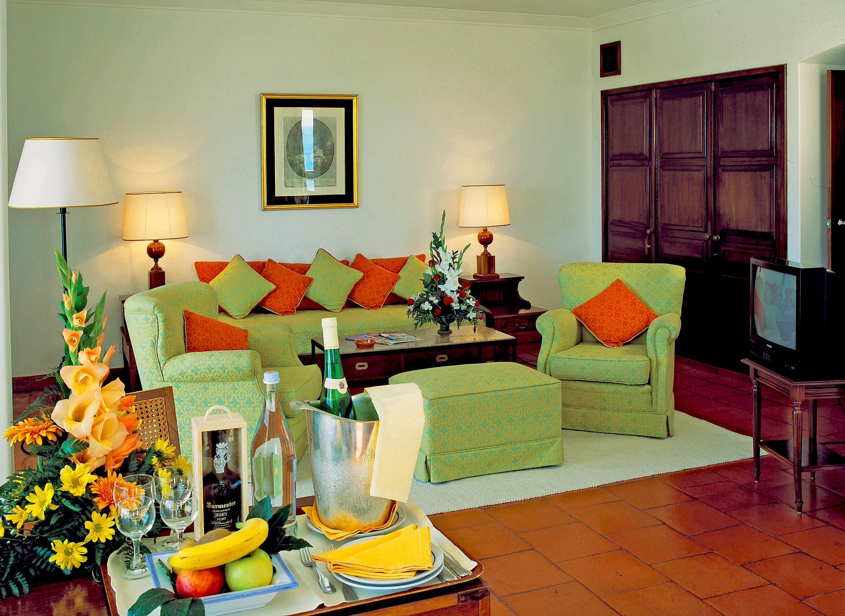 A living room with a sofa, armchairs, a coffee table with fruit and drinks, a TV, and a framed picture on the wall.