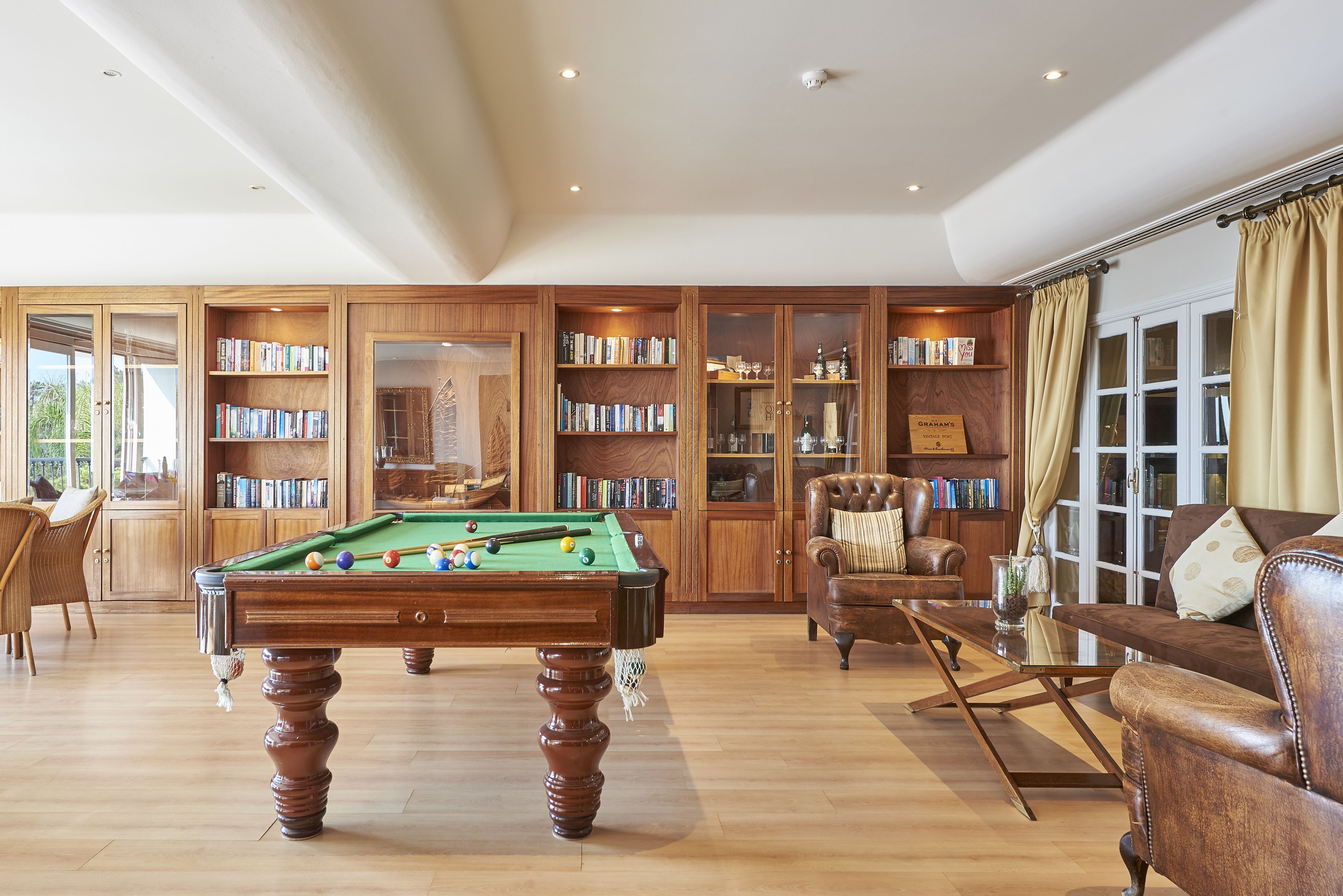 Una habitación amplia con mesa de billar, sillones de cuero y una estantería llena de libros.