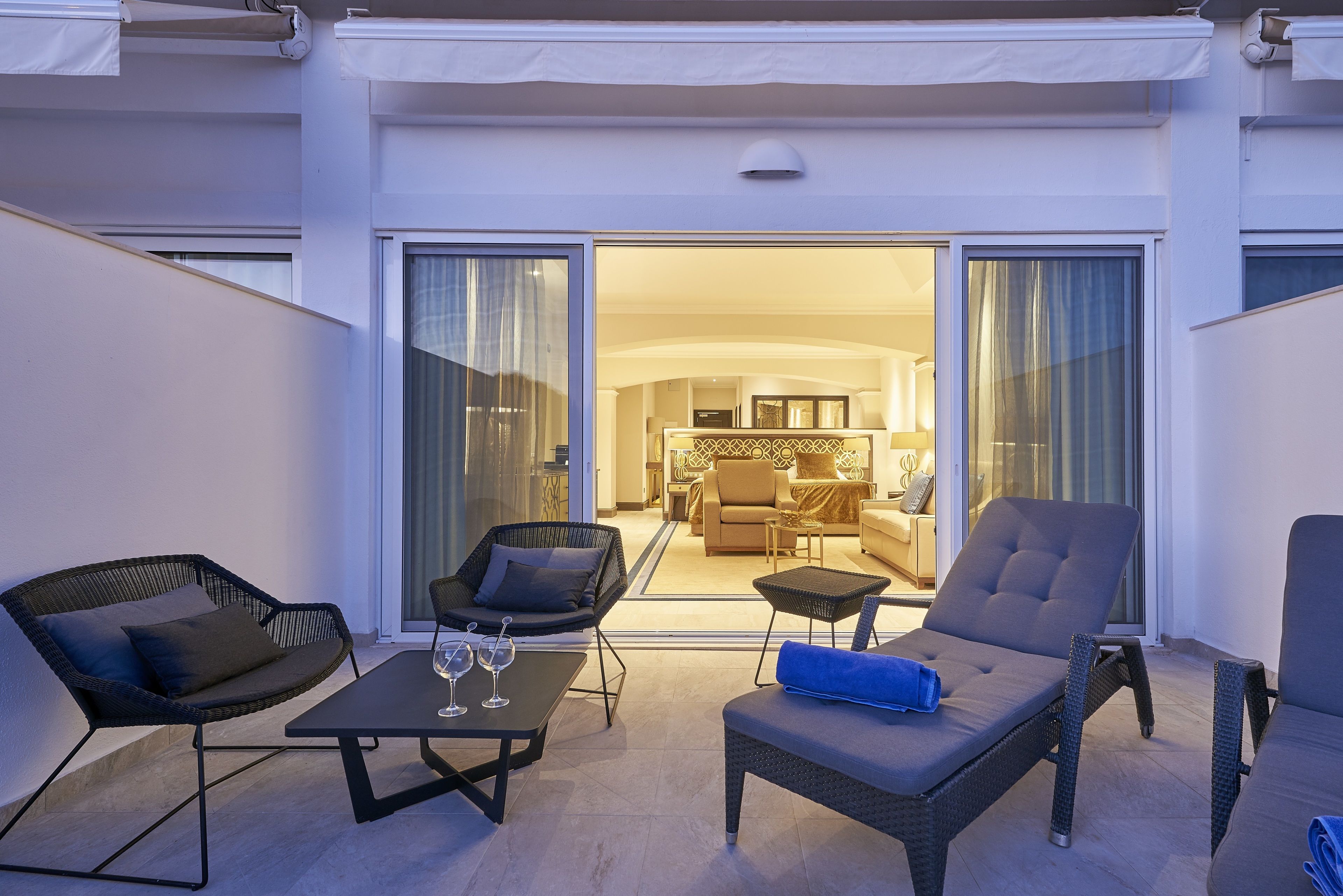 A balcony with wicker furniture, a glass table with wine glasses, and a view into a well-lit interior space.