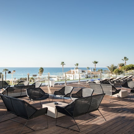 A deck with wicker chairs and a table, overlooking a coastal area with palm trees and a clear sky.