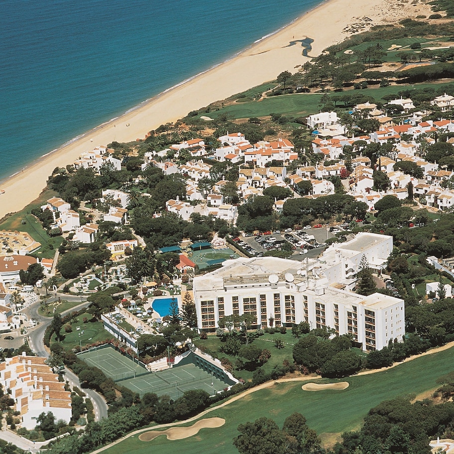 An aerial view of a coastal resort with a beach, tennis courts, and a large building with multiple floors.
