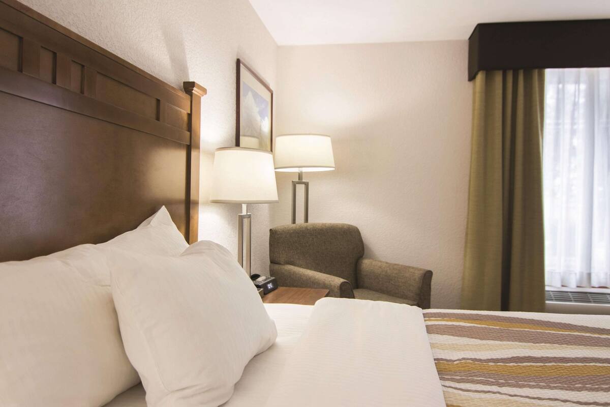 A hotel room with a large bed, a chair, a desk with a lamp, and a window with curtains.