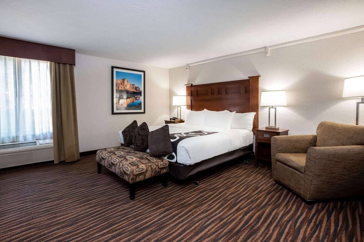 A hotel room with a bed, a chair, a small table, a picture on the wall, and a window with curtains.
