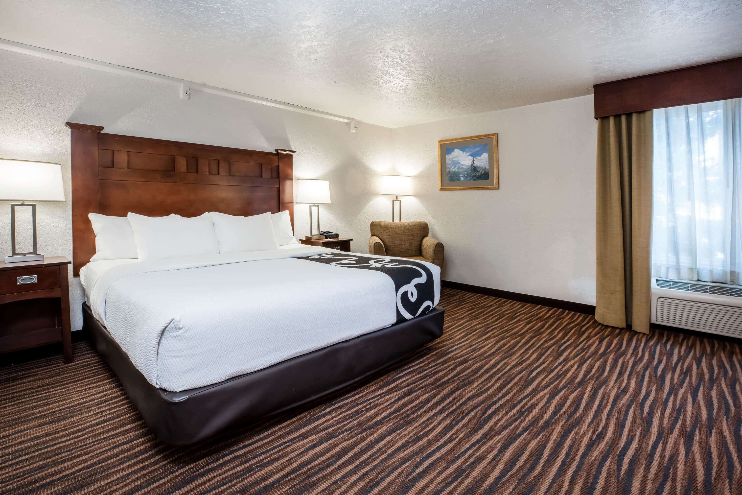 A hotel room with a large bed, a wooden headboard, a chair, two bedside lamps, a painting on the wall, and a window with curtains.