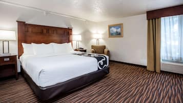 A hotel room with a large bed, a wooden headboard, a chair, two bedside lamps, a painting on the wall, and a window with curtains.