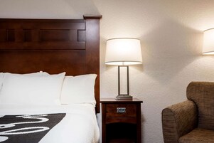 Uma cama bem arrumada com roupa de cama branca, cabeceira de madeira, abajur, mesa de cabeceira pequena de madeira com gaveta e uma poltrona estofada marrom.