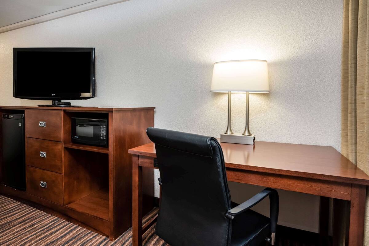 A hotel room with a wooden desk, a black office chair, a flat-screen TV, a microwave, a lamp, and a minibar.