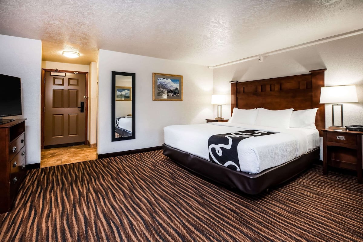 A hotel room with a large bed, a wooden headboard, two bedside tables with lamps, a flat-screen TV, a door leading to another room, and a framed picture on the wall.