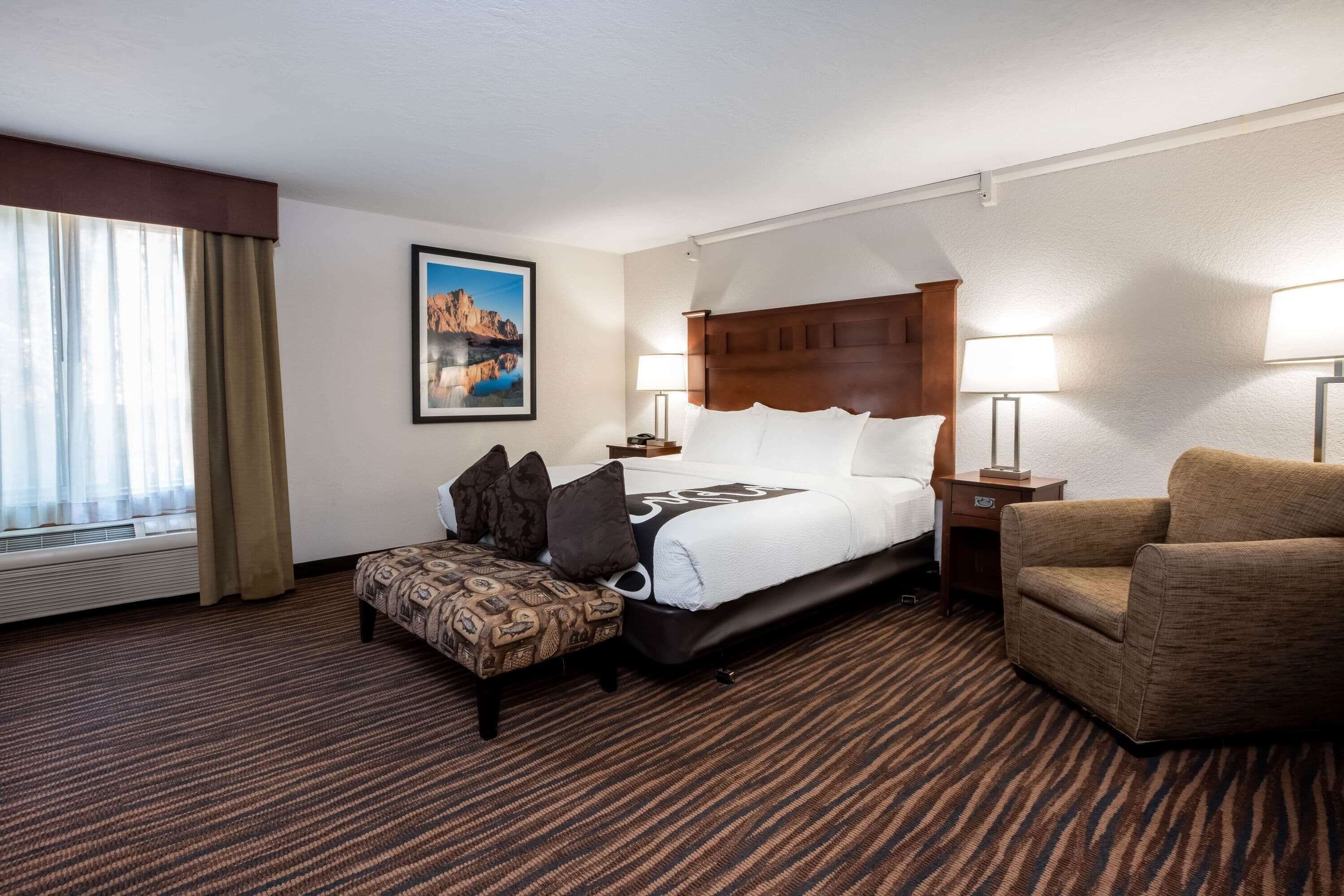 A hotel room with a bed, a chair, a small table, and a window with curtains.