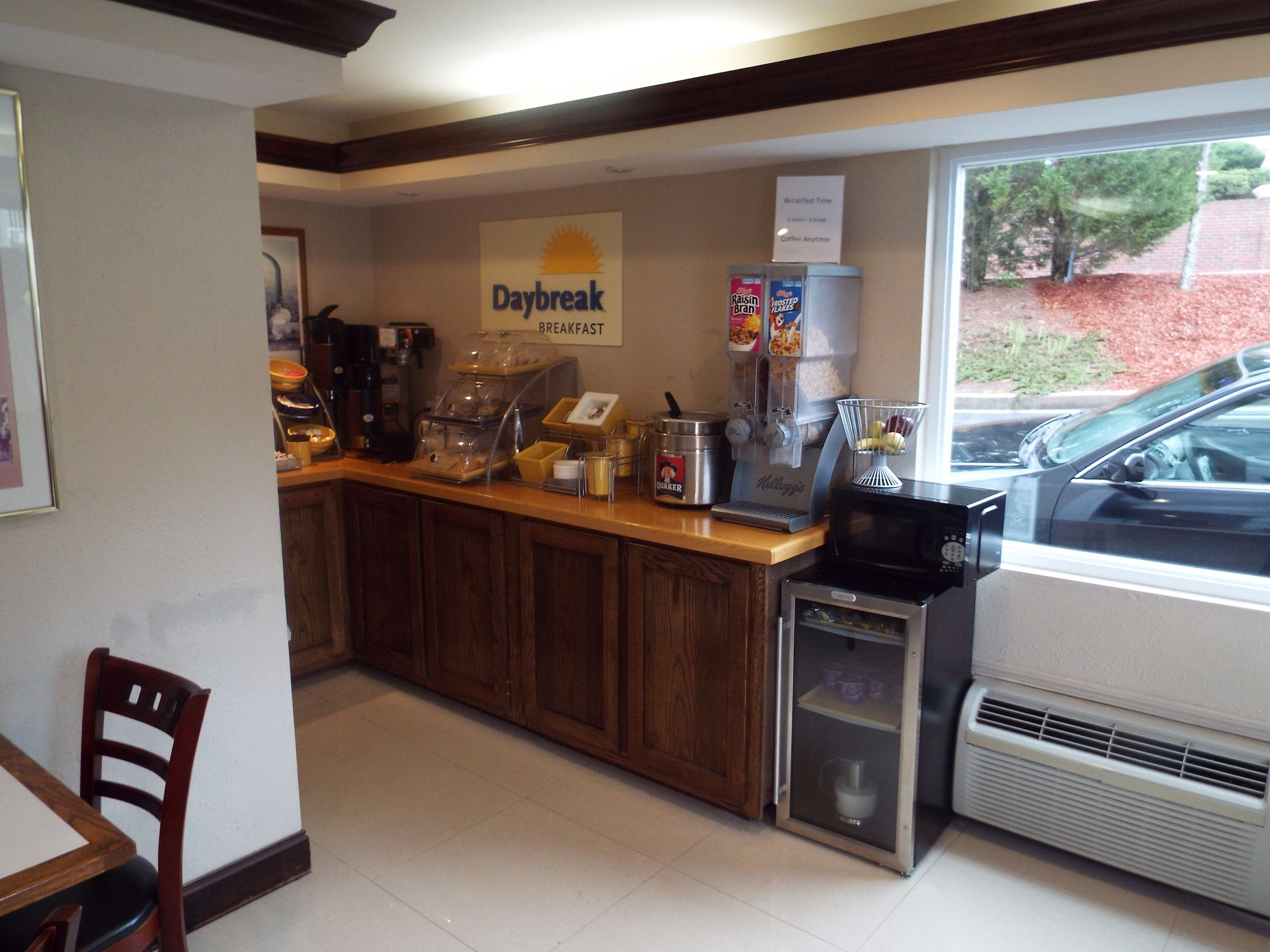 A breakfast area with a sign reading "Daybreak Breakfast," coffee machines, a small refrigerator, and a window with a view of a parked car and trees outside.