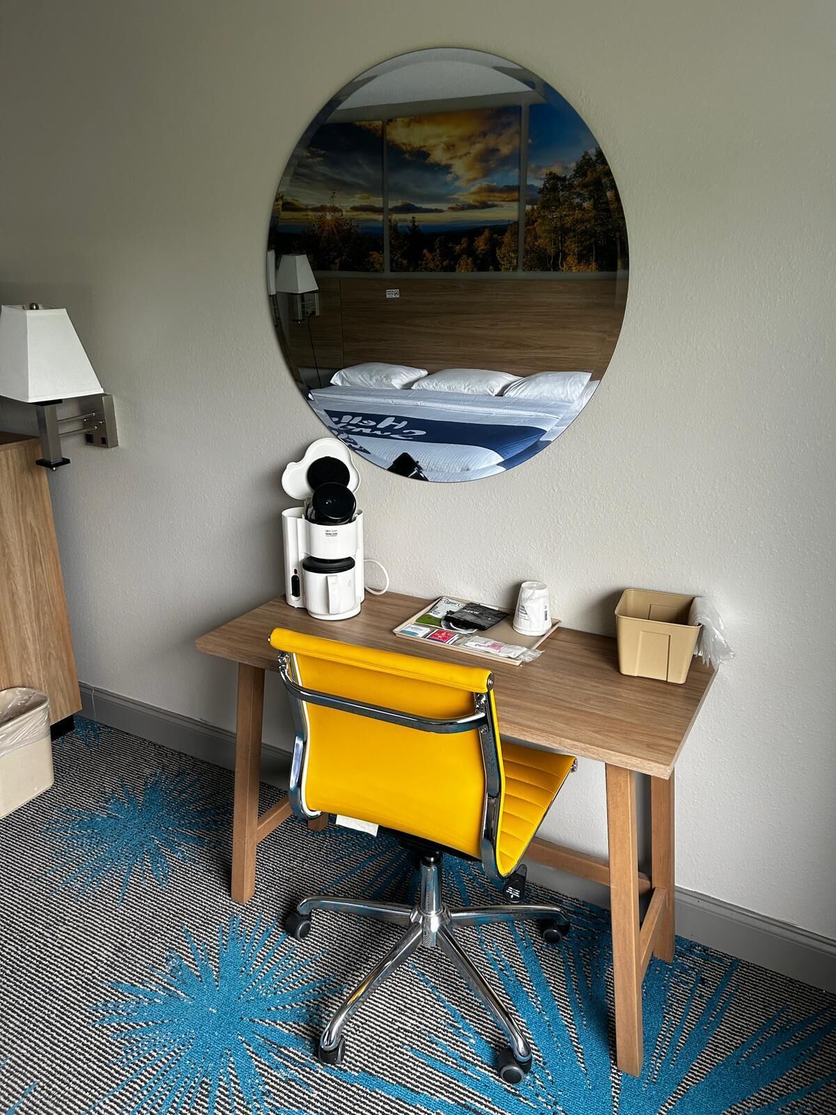 Habitación de hotel con un escritorio de madera, una silla amarilla, una cafetera, una taza, una caja de pañuelos y un espejo grande que refleja una cama y vistas a un atardecer.