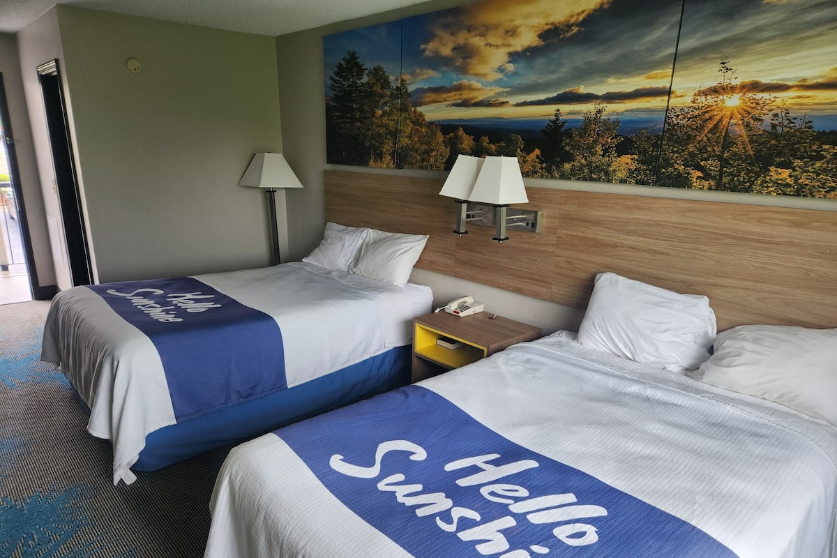 A hotel room with two beds, each with a 'Hello Sunshine' bedspread, a nightstand with a lamp, and a painting of a sunset landscape on the wall.