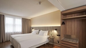 A hotel room with a large bed, a bedside table with a vase, a wooden wardrobe, and a large window with curtains. - NH Frankfurt Airport (Kelsterbach)