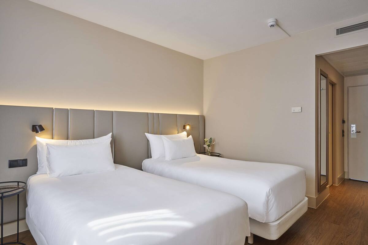 A hotel room with two beds, a bedside table with a lamp, a wall-mounted light, and a door leading to another room.