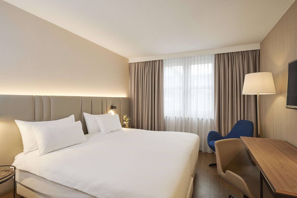 A hotel room with a large bed, a desk, a chair, a TV, and a window with curtains.