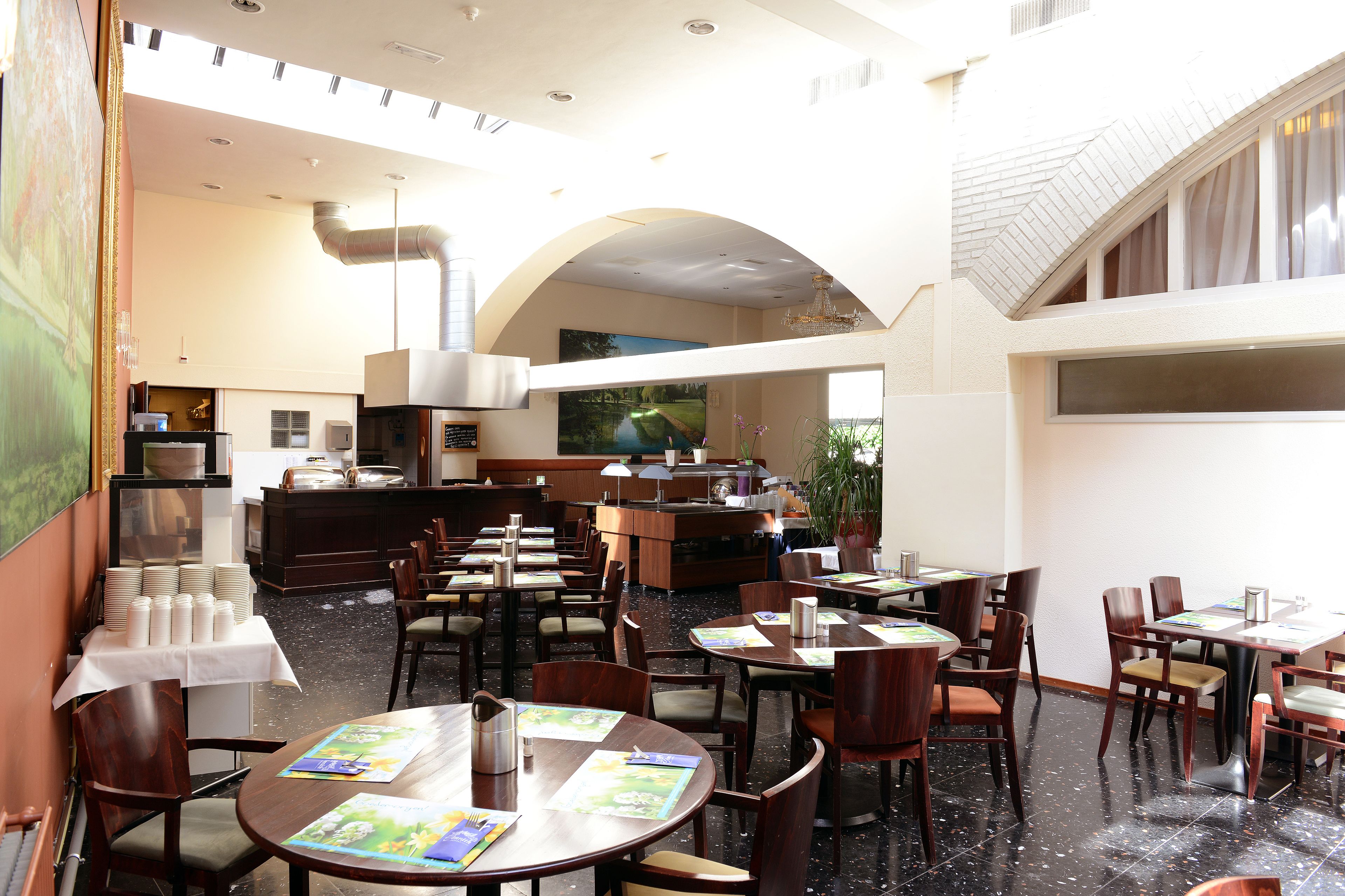 A spacious hotel lobby with multiple dining tables and chairs, a reception desk, and a large painting on the wall.