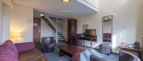 A hotel room with a purple sofa, a wooden coffee table, a desk with a chair, a mirror, and a staircase leading to another room.